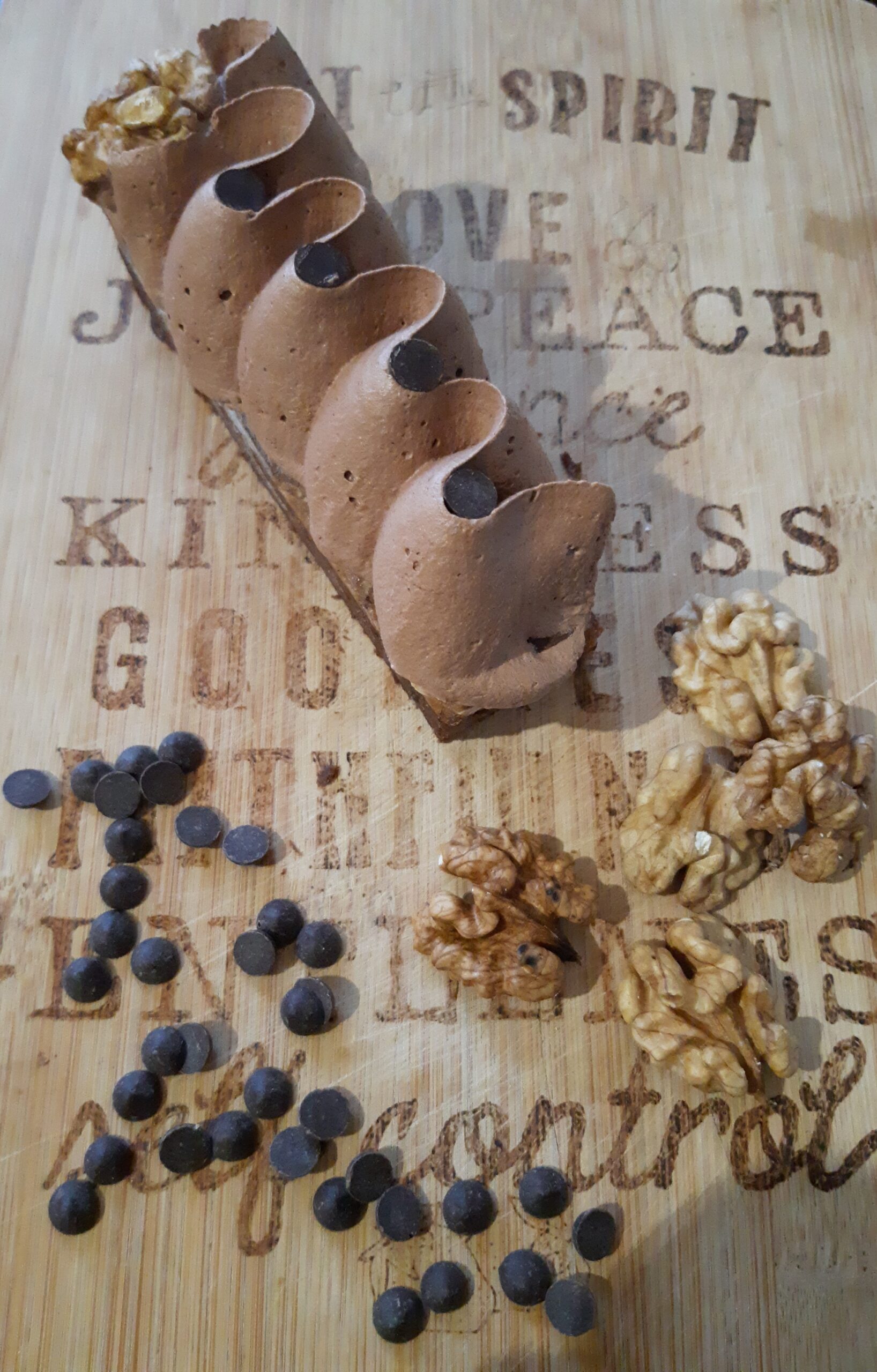 Gâteau en forme de finger, surmonté d'une ganache décorative, posée sur une planche à découpée gravée, avec quelques cerneaux de noix et des pépites de chocolat en déco
