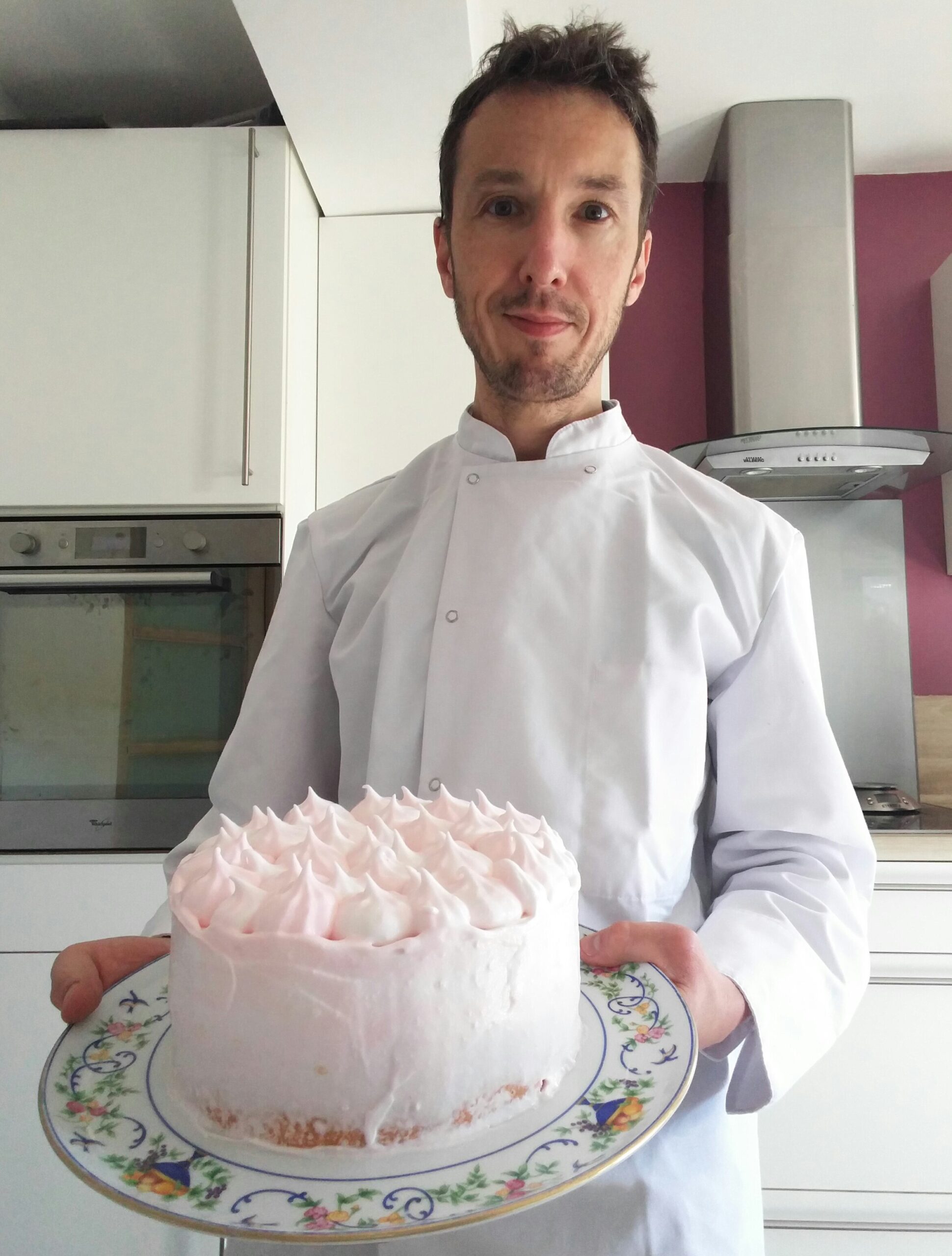 Photo de moi, en tenue de pâtissier dans ma cuisine, tenant un plat sur lequel est posé un grand bavarois blanc et rose, surmonté de pointes de chantilly rose.