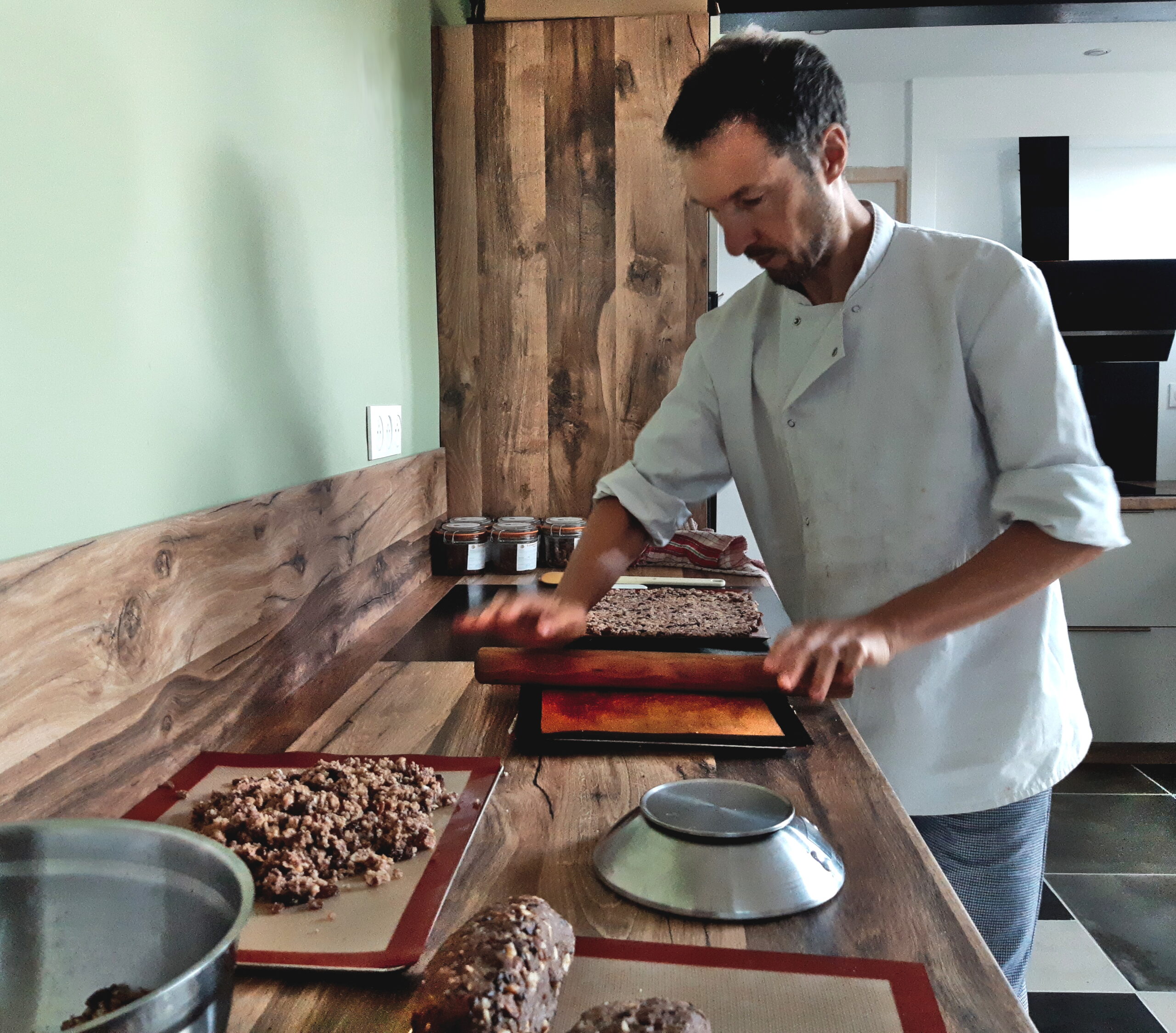 Photo de moi, en tenue de pâtissier, travaillant dans ma cuisine. Sur le plan de travail en bois, on aperçoit une balance, un cul-de-poule, et de nombreuses préparations. Je suis en train d'abaisser du greenola entre deux feuilles de silicone, avec un rouleau de pâtissier.