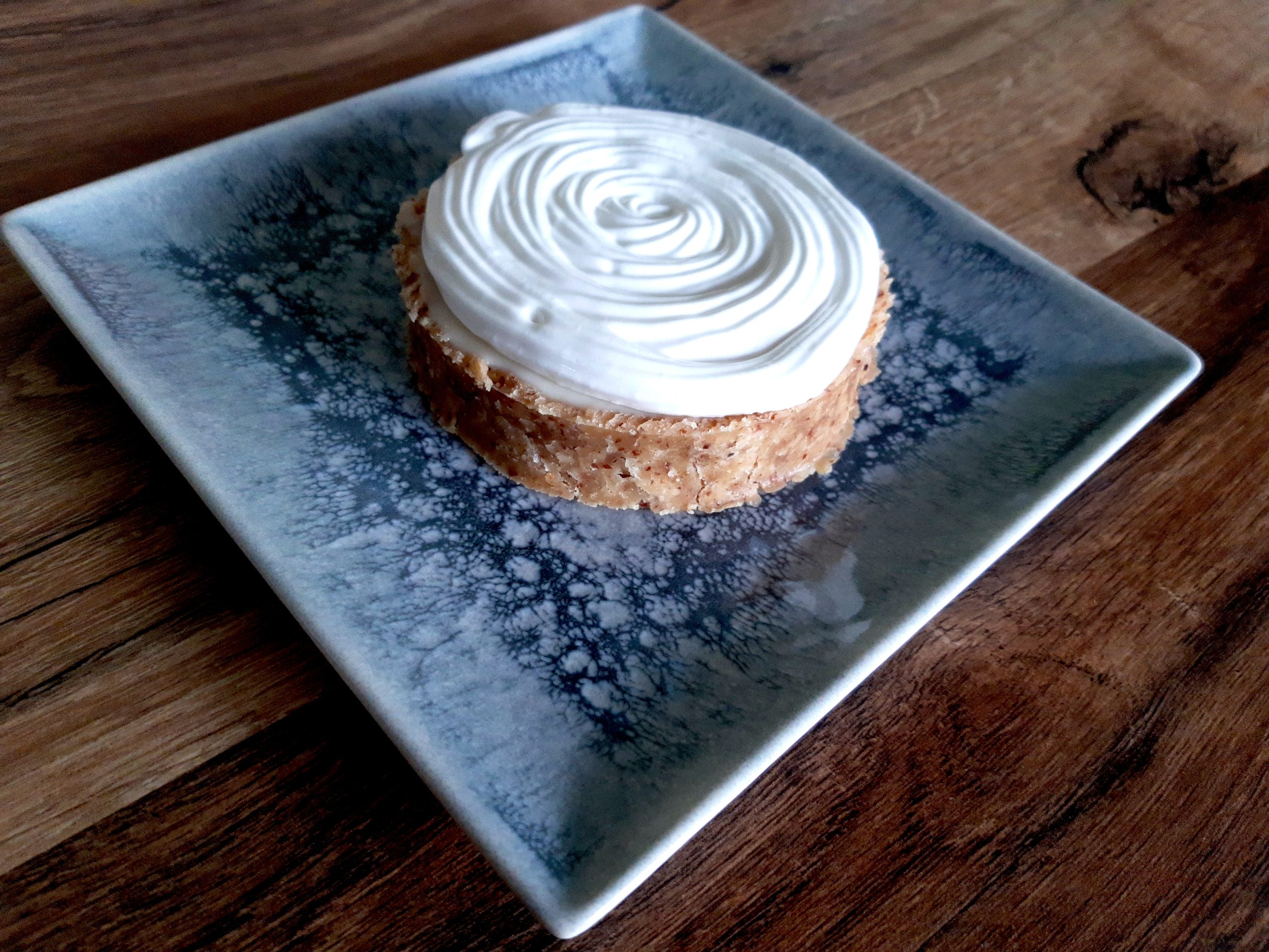 Sur une assiette posée sur un support en bois, une tartelette sans gluten garnie à la crème végétale au citron, surmontée d'une meringue italienne végétale pochée en cercles