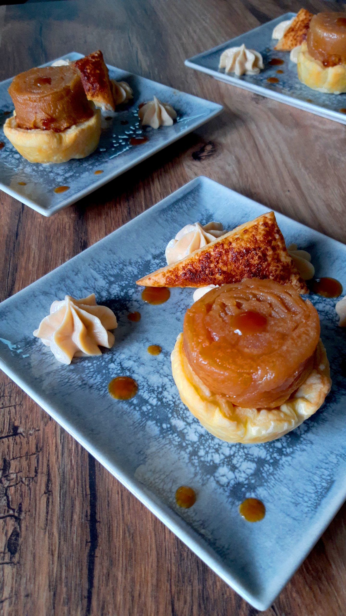 Trois assiettes contenant chacune une tarte de pâte feuilletée surmontée d'une rose de pommes compotées dans de l'huile d'olive. Autour de chaque tarte, des petites fleurs de chantilly végétale au caramel, des pointes de caramel, et un triangle de pâte feuilletée légèrement caramélisé